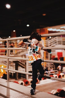 A child wearing a colorful shirt and a face mask stands playfully leaning against a metal railing in an indoor setting. The background shows an industrial-style environment with stairs and circular elements, illuminated by artificial lights.