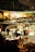 A person is sitting alone at a wooden table, writing in a notebook under warm ambient lighting. Behind them, a blurry background reveals a bustling indoor area with plants, other tables and chairs, and a bright, illuminated sign reading 'PORTER.' The atmosphere suggests a cozy, quiet spot within a vibrant café or restaurant setting.
