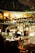 A person is sitting alone at a wooden table, writing in a notebook under warm ambient lighting. Behind them, a blurry background reveals a bustling indoor area with plants, other tables and chairs, and a bright, illuminated sign reading 'PORTER.' The atmosphere suggests a cozy, quiet spot within a vibrant café or restaurant setting.