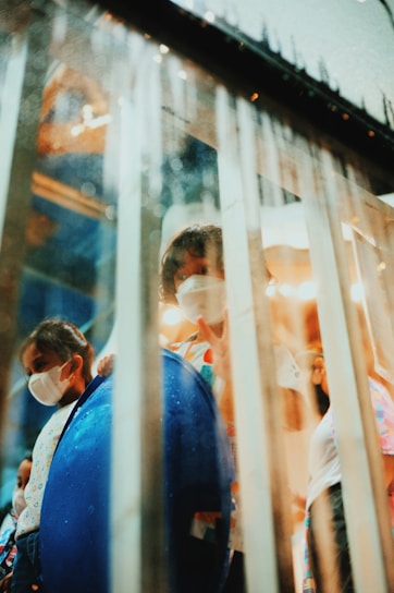 Children wearing face masks are visible behind a glass partition with droplets. One child appears to be holding up a peace sign, adding a sense of playfulness. The ambient lighting creates a soft, warm atmosphere.