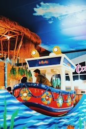 A colorful indoor amusement ride shaped like a small cartoon boat. The boat is adorned with bright red and blue colors and detailed with lifesaver rings. It is part of a playful setting with a background of faux thatched roofs and hanging greenery, and overhead is a ceiling painted like a sky with clouds.