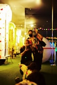 In a vibrant indoor setting, two people are positioned on a patch of green artificial turf. One person is crouching down, holding a camera, while the other stands next to them wearing a mask and making a peace sign. The background features colorful lights, reflections from mirrors, and a structure decorated with heart shapes.