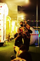 In a vibrant indoor setting, two people are positioned on a patch of green artificial turf. One person is crouching down, holding a camera, while the other stands next to them wearing a mask and making a peace sign. The background features colorful lights, reflections from mirrors, and a structure decorated with heart shapes.