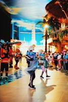 A lively indoor scene with performers in colorful costumes dancing energetically. The performers wear outfits that incorporate bright blue and black colors, with lights enhancing their appearance. They dance in a space that appears to be a themed venue, featuring palm trees and a vivid sky-painted ceiling. Spectators surround the performance area, watching the event unfold.