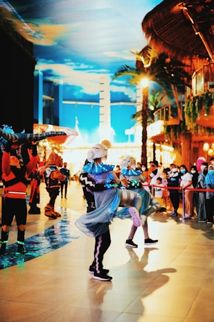 A lively indoor scene with performers in colorful costumes dancing energetically. The performers wear outfits that incorporate bright blue and black colors, with lights enhancing their appearance. They dance in a space that appears to be a themed venue, featuring palm trees and a vivid sky-painted ceiling. Spectators surround the performance area, watching the event unfold.