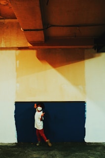 A child wearing a white face mask and a white shirt, paired with red pants, poses playfully in front of a two-toned wall. The wall is divided with an upper portion painted beige and a lower portion in dark blue. The child is holding an orange toy or object in each hand, standing on a dark surface under warm lighting.