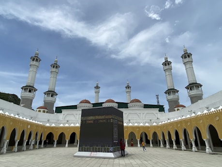 A large courtyard with an Islamic architectural style is surrounded by multiple tall white minarets and a series of arches with a golden-yellow hue. In the center of the courtyard, there is a black, cube-like structure resembling the Kaaba. People are walking around the area under a bright, partly cloudy sky.