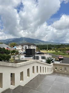 A large cumulus cloud dominates the sky with a backdrop of distant mountains. Below, a residential building is visible, surrounded by lush greenery. A staircase leads down to a courtyard area where a black monument or structure is positioned. Cars are parked nearby, and a red vehicle is seen on the road.