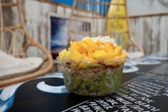 A clear plastic bowl filled with a vibrant acai bowl topped with diced mango, granola, and coconut flakes. The background features a coastal-themed decor with wicker furniture and a surfboard, creating a relaxed, beachy atmosphere.