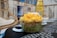 A clear plastic bowl filled with a vibrant acai bowl topped with diced mango, granola, and coconut flakes. The background features a coastal-themed decor with wicker furniture and a surfboard, creating a relaxed, beachy atmosphere.