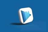 A 3D icon with a play button symbol and a book graphic is centered against a solid blue background. The icon is designed in white and shades of blue, with a modern and sleek appearance.