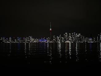 A nighttime skyline with a cityscape illuminated by various colorful lights. The prominent CN Tower is lit with green, red, and white colors. The reflection of the lights shimmers on the water in the foreground, and a stadium with a glowing dome is visible.