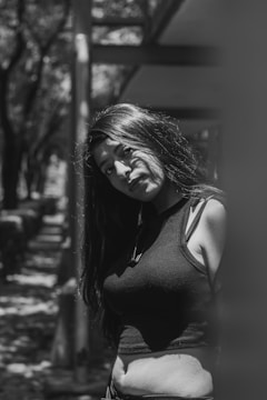 A black and white image of a woman with long hair leaning slightly against a wall. She is wearing a sleeveless top and looking towards the camera. The lighting creates contrasting shadows on her face and the surrounding environment. The background includes blurred trees and architectural elements, forming a natural, contemplative scene.