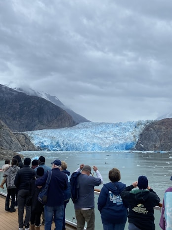 A group of people stand on a viewing platform, dressed in warm clothing, observing a massive glacier with a serene body of water in front. The glacier is nestled between rocky mountains under an overcast sky.