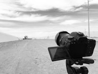 A DSLR camera is set on a tripod, pointing towards an open landscape with a dirt road stretching into the distance. The sky is partially cloudy, and there are some structures barely visible on the horizon.
