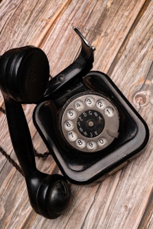 A vintage black rotary dial telephone with a coiled cord is placed on a wooden surface. The phone features a circular dial with numbers and letters, and the handset rests off the hook next to the base.