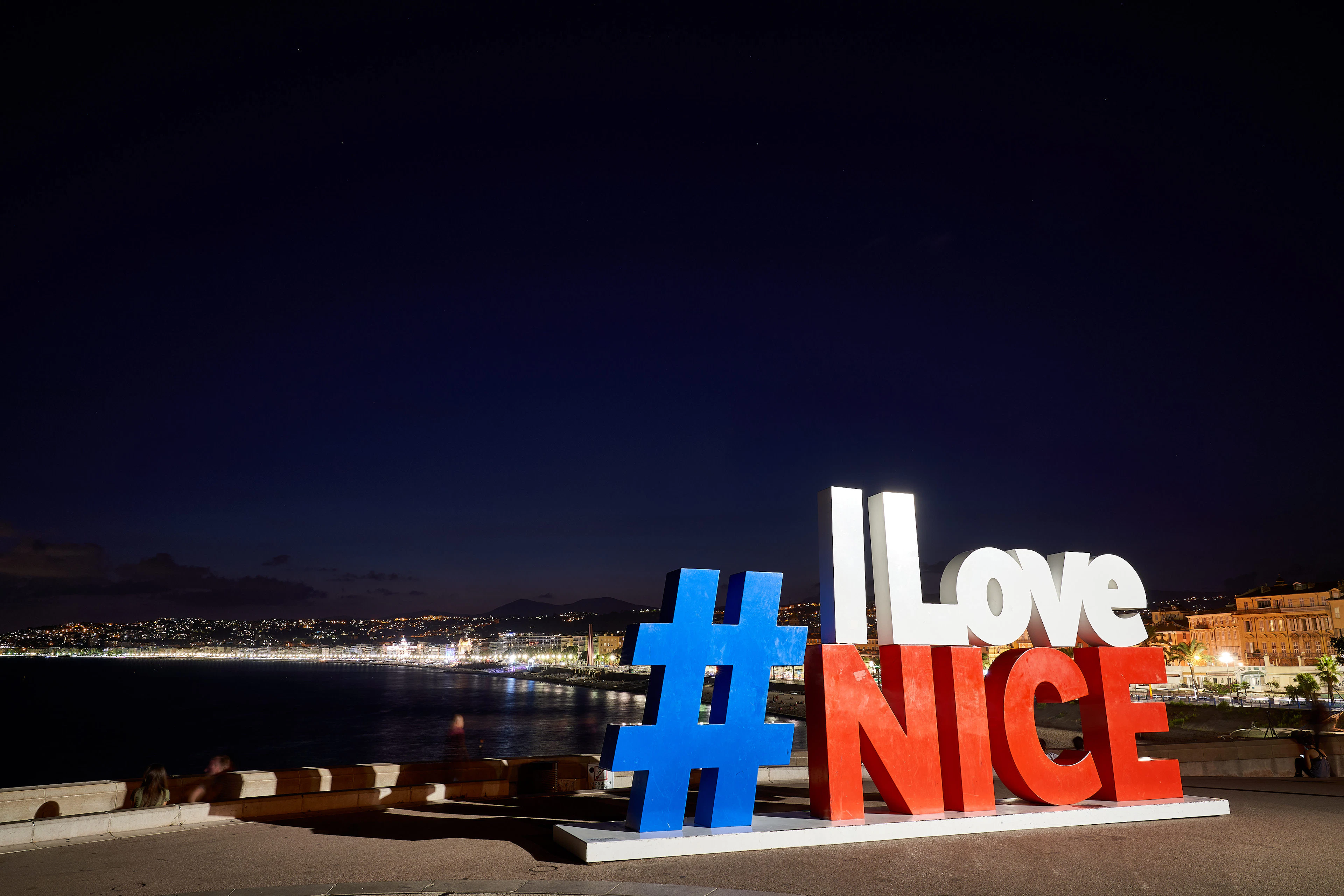 The illuminated 'I Love Nice' sign glows at night above the bay under a starry sky.