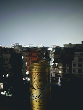 A golden can with intricate designs is prominently displayed against a blurred nighttime cityscape. The background features dimly lit buildings and a cloudy sky, creating a contrast with the can's reflective surface.