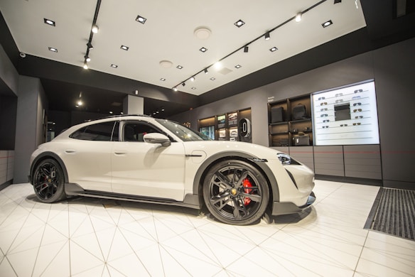 A sleek, white sports car is prominently displayed in a modern, upscale showroom. The environment is well-lit, with spotlights emphasizing the car’s design. Shelving units in the background showcase various items, adding a luxurious feel to the space.