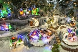 A charming miniature Christmas village scene with snow-covered streets and buildings, surrounded by decorated pine trees. Various small figures are engaged in festive activities, illuminated by multicolored lights. The setting includes houses, small businesses, and trees adorned with snow, creating a cozy and festive atmosphere.