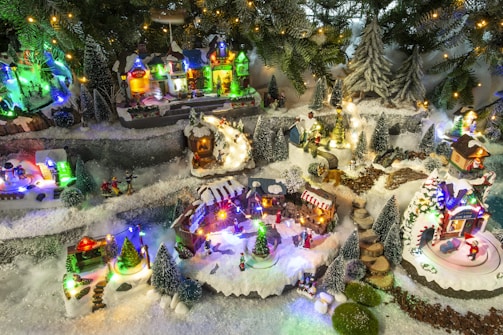 A charming miniature Christmas village scene with snow-covered streets and buildings, surrounded by decorated pine trees. Various small figures are engaged in festive activities, illuminated by multicolored lights. The setting includes houses, small businesses, and trees adorned with snow, creating a cozy and festive atmosphere.