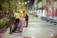 Two workers in uniform are collecting trash at night on a city street. They are using a large bag to gather waste, near a line of empty benches. The street is illuminated with artificial lights and lined with trees, creating an urban yet serene atmosphere.