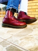 A pair of shiny, dark red boots with yellow stitching are worn with faded blue jeans on a tiled floor. The jeans have frayed hems, adding a casual touch to the overall appearance. The background features a tan brick wall and a window with a view of the outdoors.