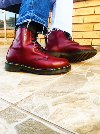 A pair of shiny, dark red boots with yellow stitching are worn with faded blue jeans on a tiled floor. The jeans have frayed hems, adding a casual touch to the overall appearance. The background features a tan brick wall and a window with a view of the outdoors.