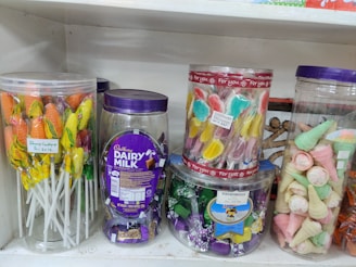 A shelf displays a variety of colorful candies in transparent plastic jars. On the left, there are lollipops with orange and yellow colors. The center jar contains Cadbury Dairy Milk chocolates, noticeable by the distinctive purple packaging. Another jar holds candies shaped like butterflies in pastel shades. On the right, wafer cones with pink and green colors are visible.