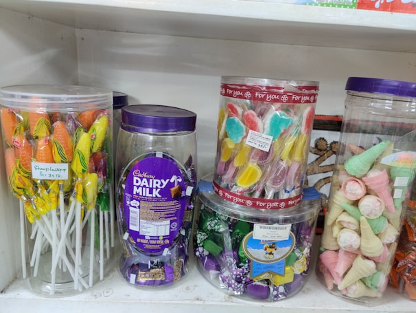 A shelf displays a variety of colorful candies in transparent plastic jars. On the left, there are lollipops with orange and yellow colors. The center jar contains Cadbury Dairy Milk chocolates, noticeable by the distinctive purple packaging. Another jar holds candies shaped like butterflies in pastel shades. On the right, wafer cones with pink and green colors are visible.