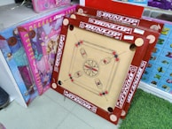 A carrom board with red borders and a wooden playing surface stands upright against a shelf. The board is surrounded by brightly colored packaging boxes featuring butterfly designs and doll images. The setting appears to be a retail space with tiled flooring and a patch of green carpet.