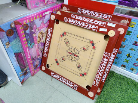 A carrom board with red borders and a wooden playing surface stands upright against a shelf. The board is surrounded by brightly colored packaging boxes featuring butterfly designs and doll images. The setting appears to be a retail space with tiled flooring and a patch of green carpet.