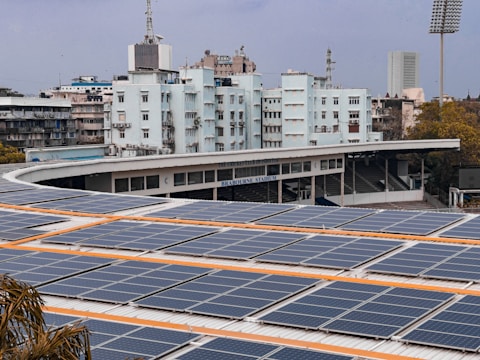 A large array of solar panels is installed on the roof of a stadium building. Behind the stadium, there are several buildings, one of which is labeled 'Brabourne Stadium'. The sky is overcast, and there are trees visible in the foreground.