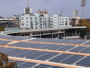 A large array of solar panels is installed on the roof of a stadium building. Behind the stadium, there are several buildings, one of which is labeled 'Brabourne Stadium'. The sky is overcast, and there are trees visible in the foreground.