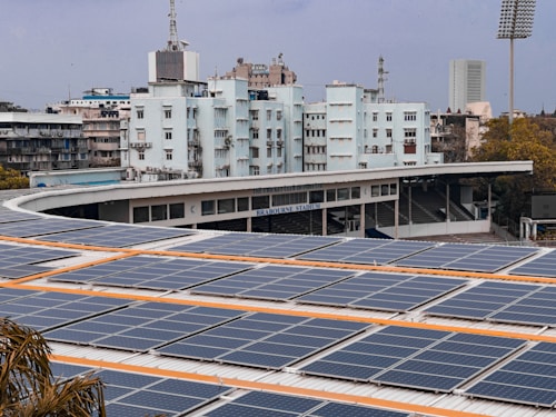 A large array of solar panels is installed on the roof of a stadium building. Behind the stadium, there are several buildings, one of which is labeled 'Brabourne Stadium'. The sky is overcast, and there are trees visible in the foreground.