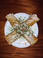 Four folded crepes are arranged in a cross pattern on a white plate. The crepes are garnished with chopped herbs, grated cheese, and small pieces of vegetables, creating a visually appealing dish.