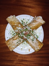 Four folded crepes are arranged in a cross pattern on a white plate. The crepes are garnished with chopped herbs, grated cheese, and small pieces of vegetables, creating a visually appealing dish.