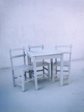 A simple, minimalist scene featuring a small white table accompanied by three matching white wooden chairs. The setting is against a pale, textured wall, creating a calming and serene environment with a monochromatic color scheme.