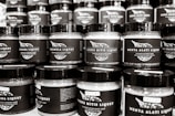 A collection of jars with black labels arranged in rows, each featuring different text and designs. The jars have a consistent shape and size with black lids, and they appear to be filled with similar contents.