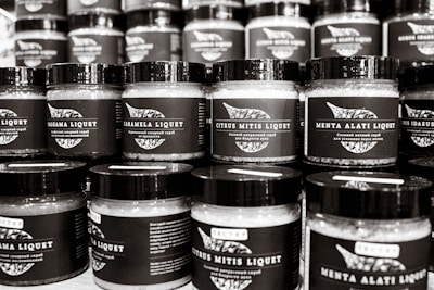 A collection of jars with black labels arranged in rows, each featuring different text and designs. The jars have a consistent shape and size with black lids, and they appear to be filled with similar contents.