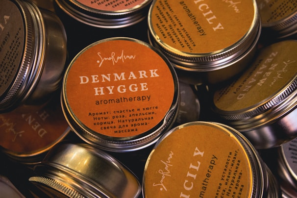 Several round, metal tins with lids featuring labels that have various aromatic scents such as Denmark Hygge and Sicily. The labels are mostly orange and beige with a mix of English and non-English languages detailing the scents.