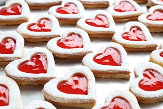 Heart-shaped cookies with a red jelly center and dusted with powdered sugar are arranged on a surface.