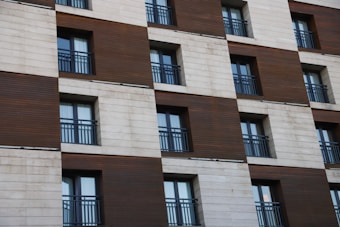 A modern building facade with alternating vertical wooden panels and pale stone or concrete sections, featuring multiple windows with dark metal railings.