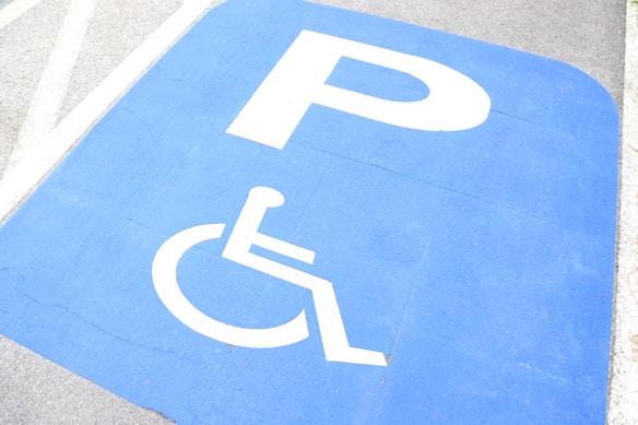 A blue parking space reserved for disabled access, marked with a white wheelchair symbol and a large 'P'. The background includes gray pavement and white lines.