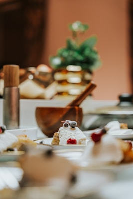 A close-up view of a collection of dessert plates featuring a small cake topped with whipped cream, berries, and sprinkled with powdered sugar. In the background, there's a blurred-out wooden mortar and pestle with green foliage, creating a cozy and inviting atmosphere.