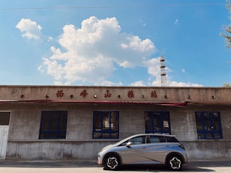 A modern car is parked in front of a rustic building with blue-framed windows and red signage. Above the building, a clear day sky filled with white clouds is visible, and a tall communication tower is seen in the background.