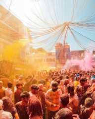 A large, vibrant crowd gathers outdoors, celebrating what appears to be a festival with colorful powders filling the air. Bright colors like yellow and pink are prominent, and people are joyfully throwing powder and interacting with each other. The atmosphere seems festive and lively.