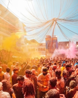 A large, vibrant crowd gathers outdoors, celebrating what appears to be a festival with colorful powders filling the air. Bright colors like yellow and pink are prominent, and people are joyfully throwing powder and interacting with each other. The atmosphere seems festive and lively.
