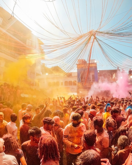 A large, vibrant crowd gathers outdoors, celebrating what appears to be a festival with colorful powders filling the air. Bright colors like yellow and pink are prominent, and people are joyfully throwing powder and interacting with each other. The atmosphere seems festive and lively.