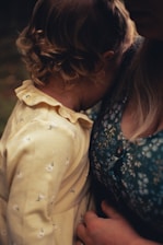 A young child with curly hair is nestled closely against an adult. The child wears a light yellow dress with small floral patterns, while the adult is in a blue patterned shirt. The scene conveys a sense of closeness and comfort.